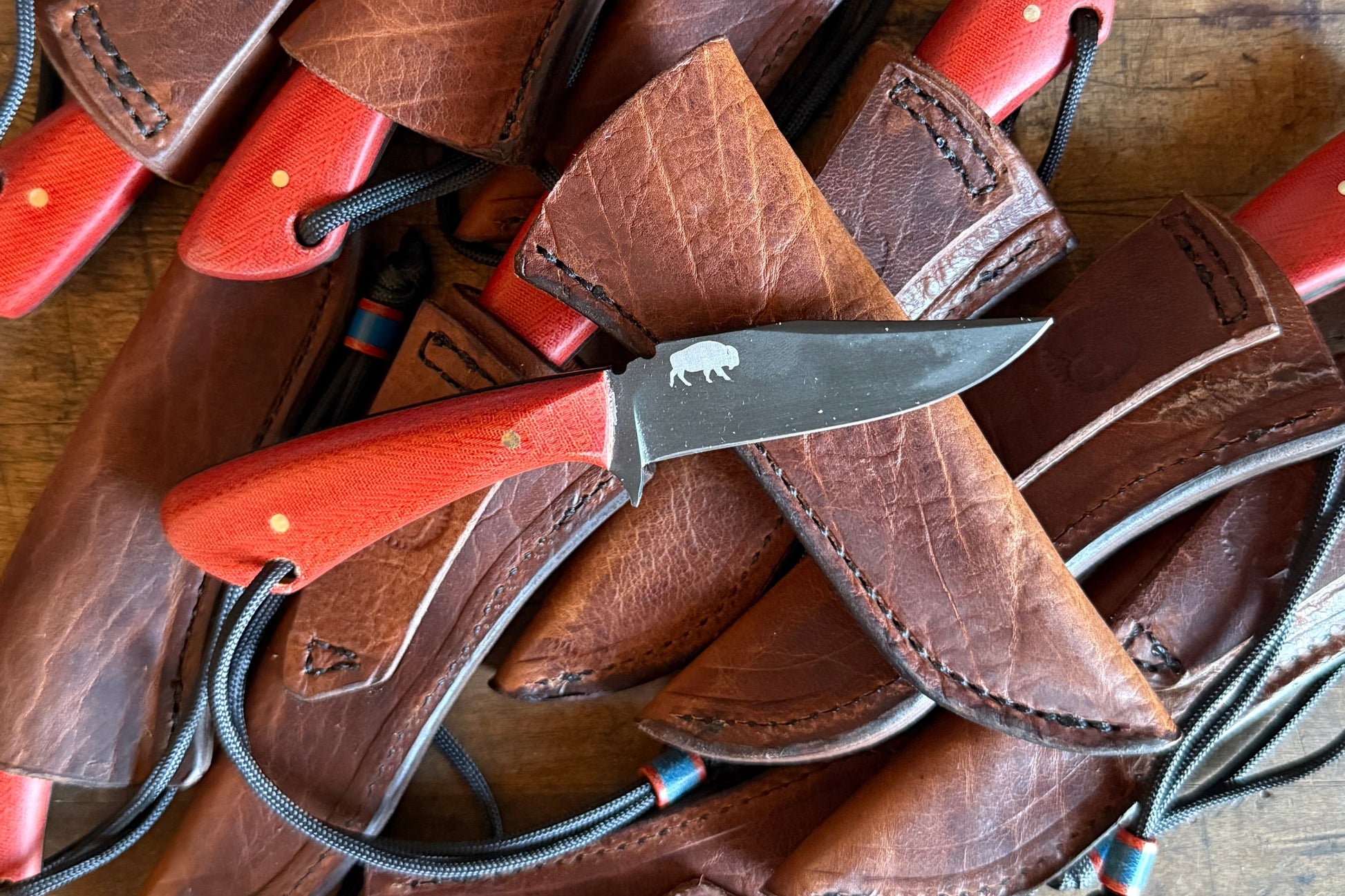 A pile of leather sheaths and knives with red handles arranged on a rustic wooden table.